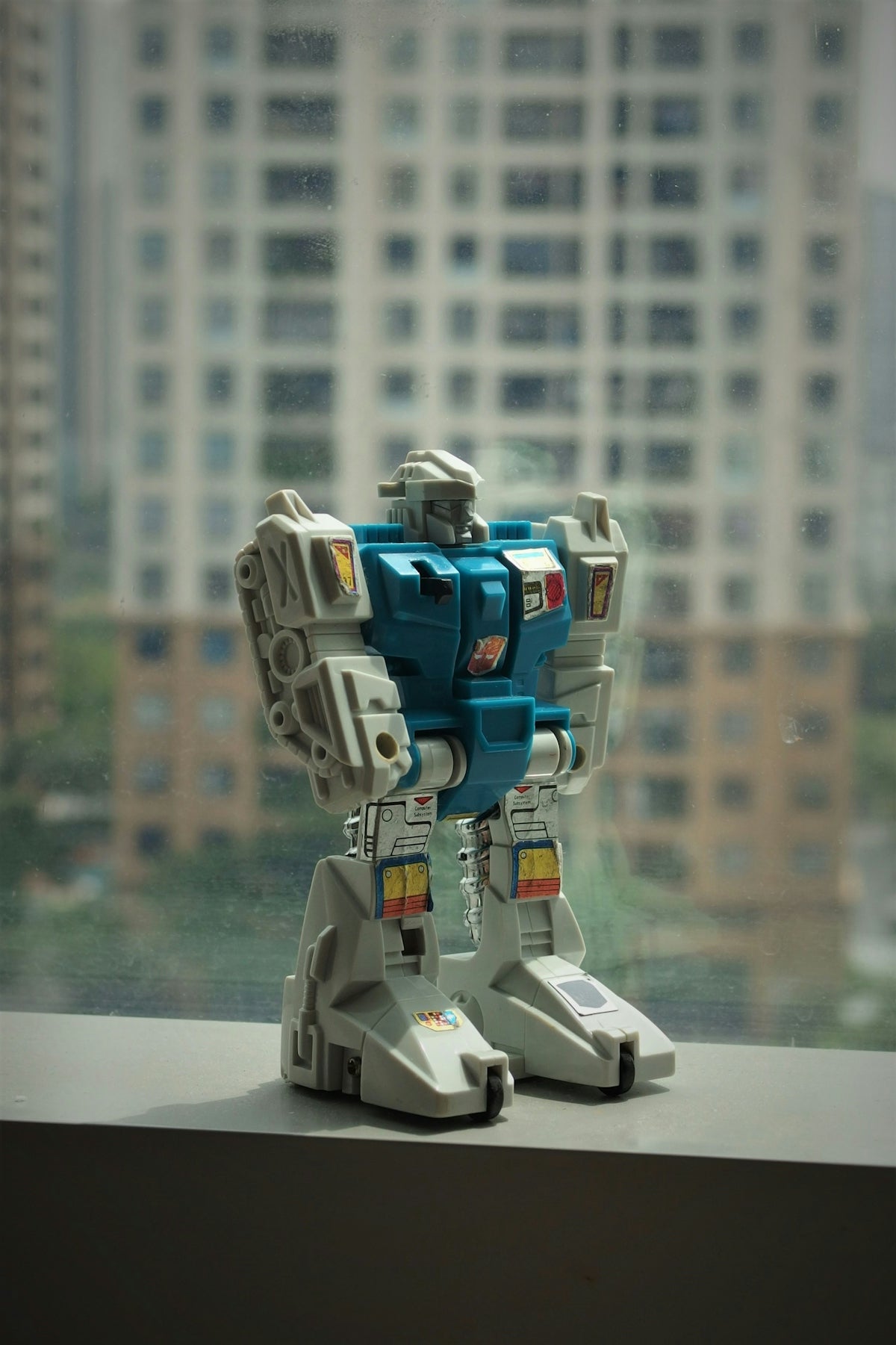 Robot toy stands on a windowsill overlooking a city.